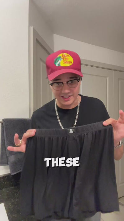 Man in glasses and red cap holding black Mr. Fertyl boxer shorts in bathroom, sharing fertility-safe underwear testimonial.